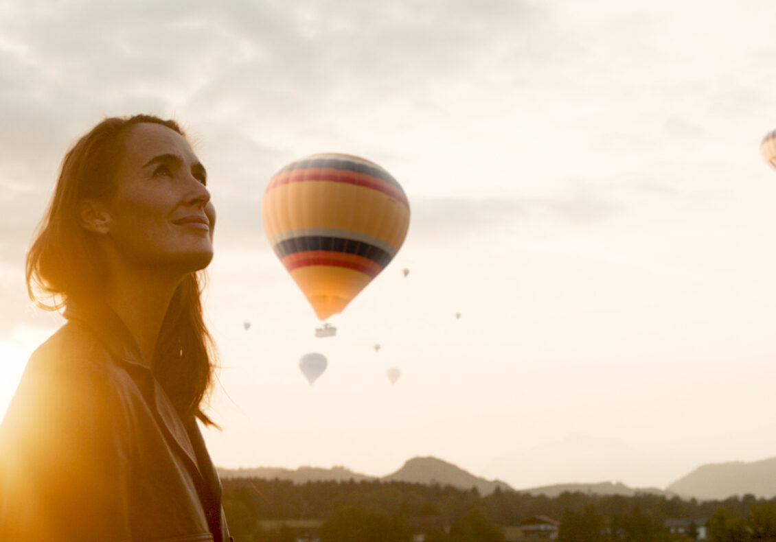 A lady looks up, smiling at hot air balloons in the glow of the evening sky in Carinthia.