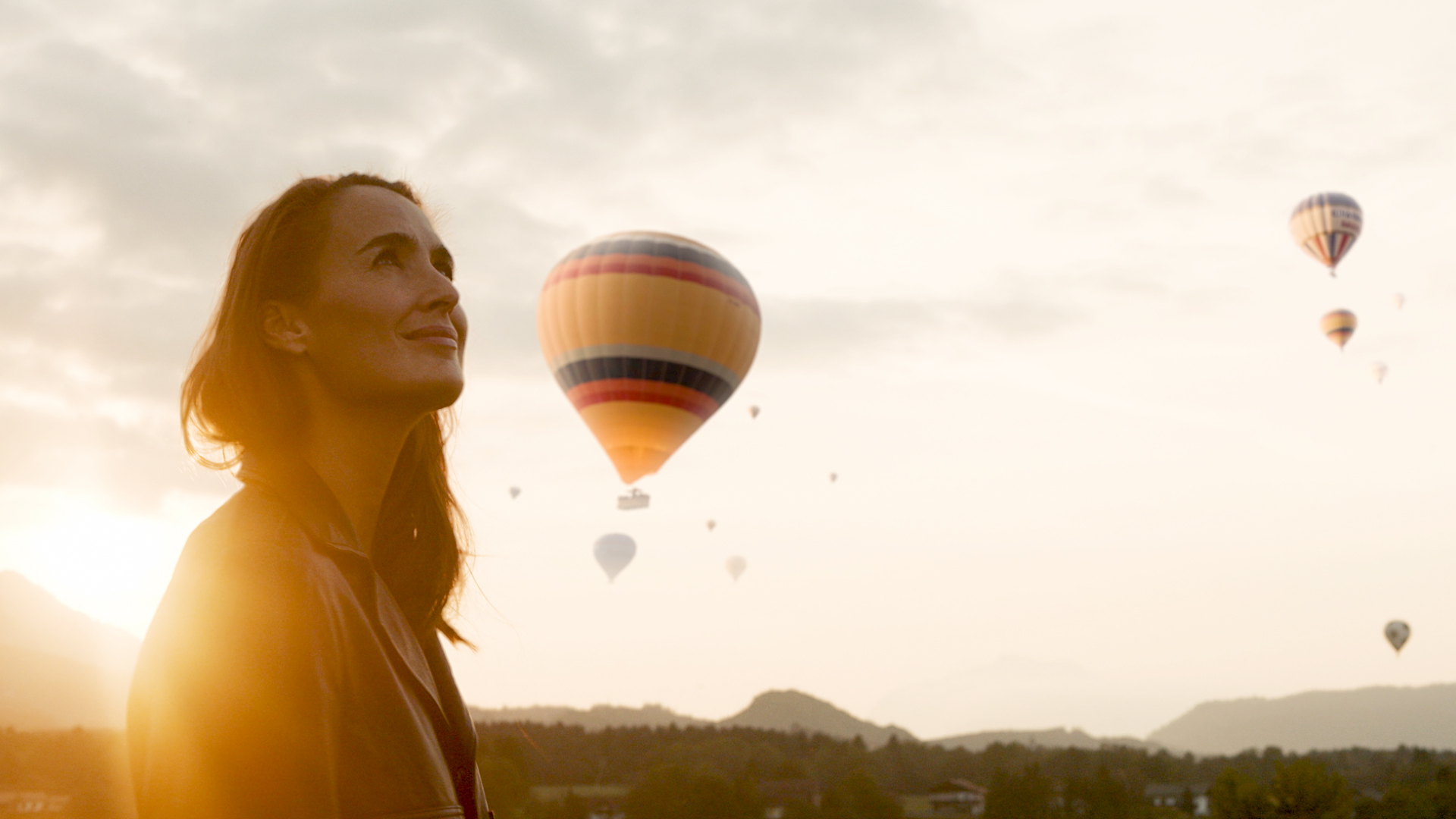 A lady looks up, smiling at hot air balloons in the glow of the evening sky in Carinthia.
